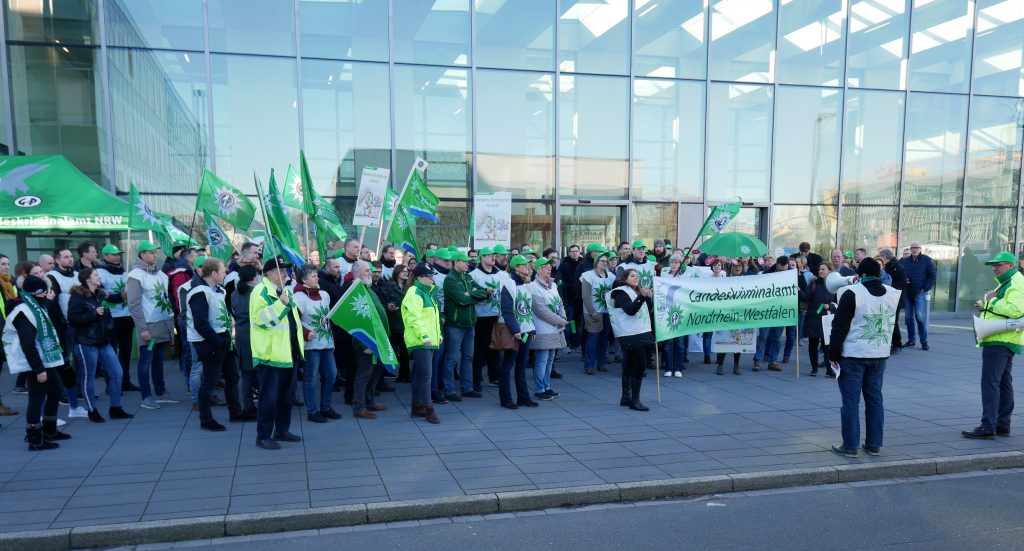 Vor dem Haupteingang des LKA NRW stehen viele Demonstranten mit leuchtenden Leibchen und Plakaten. Im Hintergrund spiegelt sich der blaue Himmel an der Glasfront des Foyers.